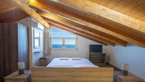 A bedroom with a wooden ceiling, a bed, a TV, and a window with a view of the sea.