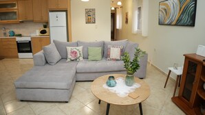 A modern living room with a grey sectional sofa, a round wooden coffee table, and a kitchen area with wooden cabinets and a refrigerator. - Nikis Country House - Lefkada BnB  Private Parking (Ligia)
