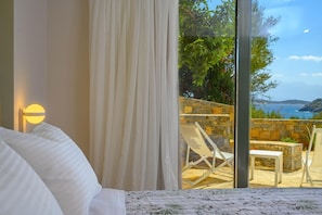 A bedroom with a bed, a large window with curtains, and a view of an outdoor seating area with chairs and a table. - Heartwood Suite w Shared Pool by Roots Suites (Agios Nikolaos)