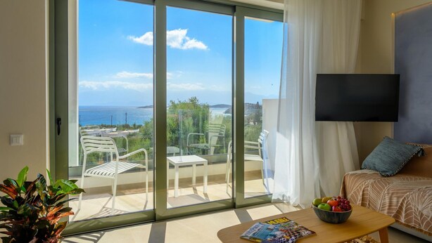 A modern room with a large sliding glass door leading to a balcony with outdoor furniture. A flat-screen TV is mounted on the wall, and a magazine is on the coffee table. - Branch Suite w Shared Pool by Roots Suites (Agios Nikolaos)
