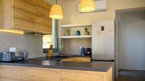 A modern kitchen with wooden cabinetry, a central island, pendant lighting, and a built-in air conditioning unit. - Branch Suite w Shared Pool by Roots Suites (Agios Nikolaos)