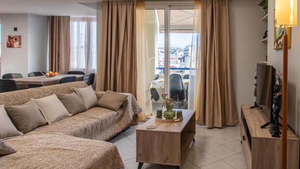 A modern living room with a large sofa, a wooden coffee table, and a TV stand. There is a balcony with a view of the city. - Sky 7 View Apartment (Heraklion)