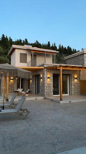 A two-story house with a stone facade, a balcony, and a covered outdoor seating area with lounge chairs.