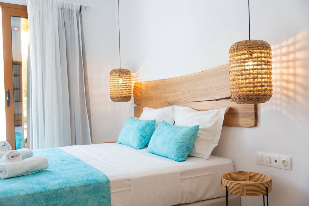 Un lit bien fait, avec une tête de lit en bois, deux oreillers bleus et une couverture blanche. Deux lampes suspendues en fibres tressées sont accrochées au-dessus du lit.