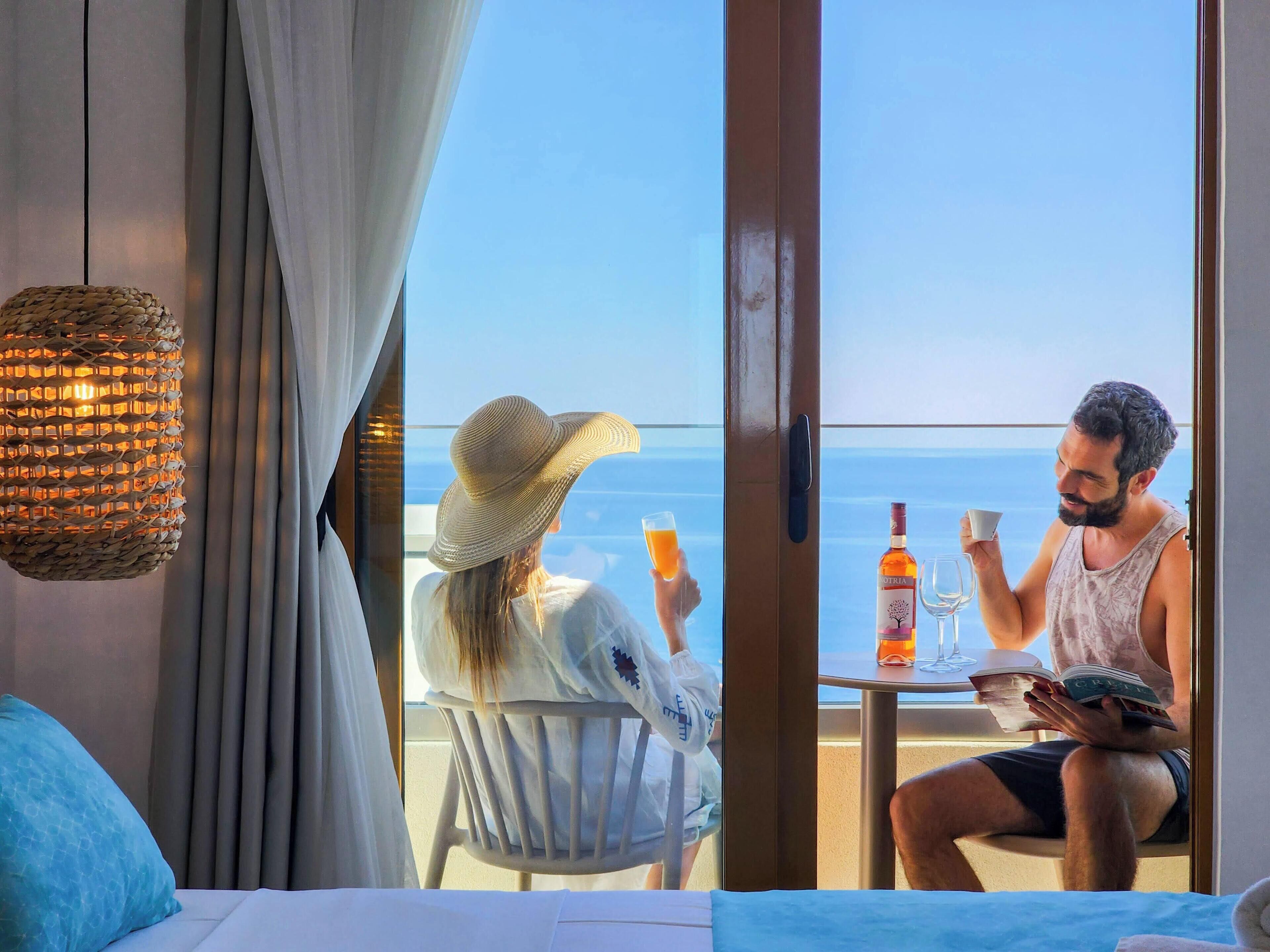 A room with a bed, a window with curtains, a wicker lamp, and a table with a bottle and glasses.