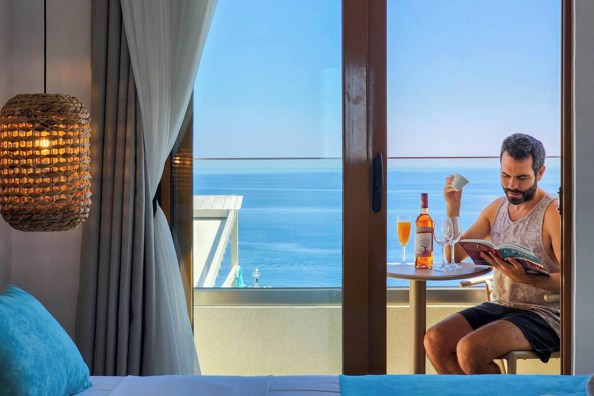 Une pièce dotée d’une grande fenêtre donnant sur la mer, d’un lit, d’une table avec une bouteille et des verres, et d’un luminaire suspendu.