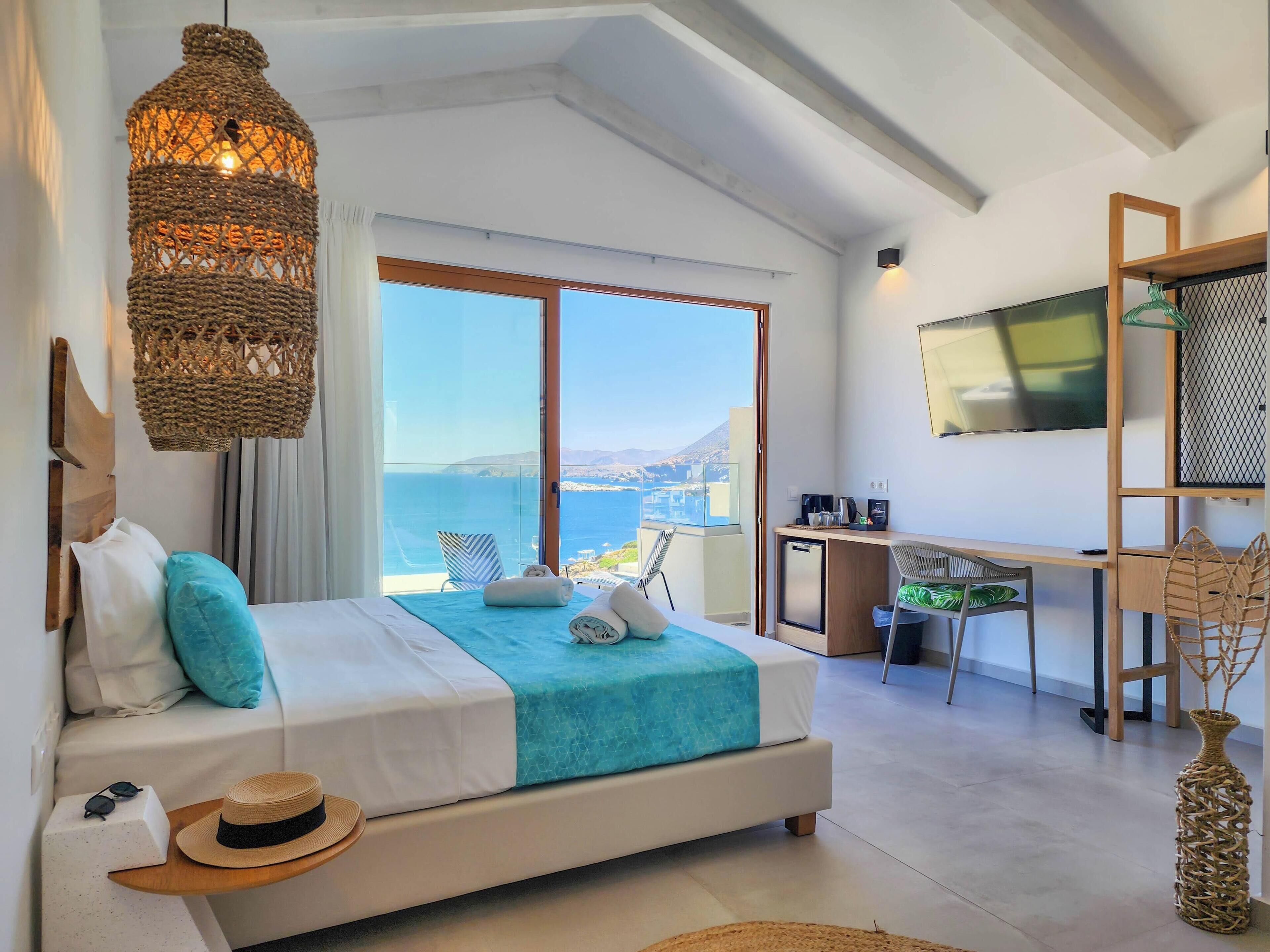 A bedroom with a bed, a desk, a TV, and a view of the sea.