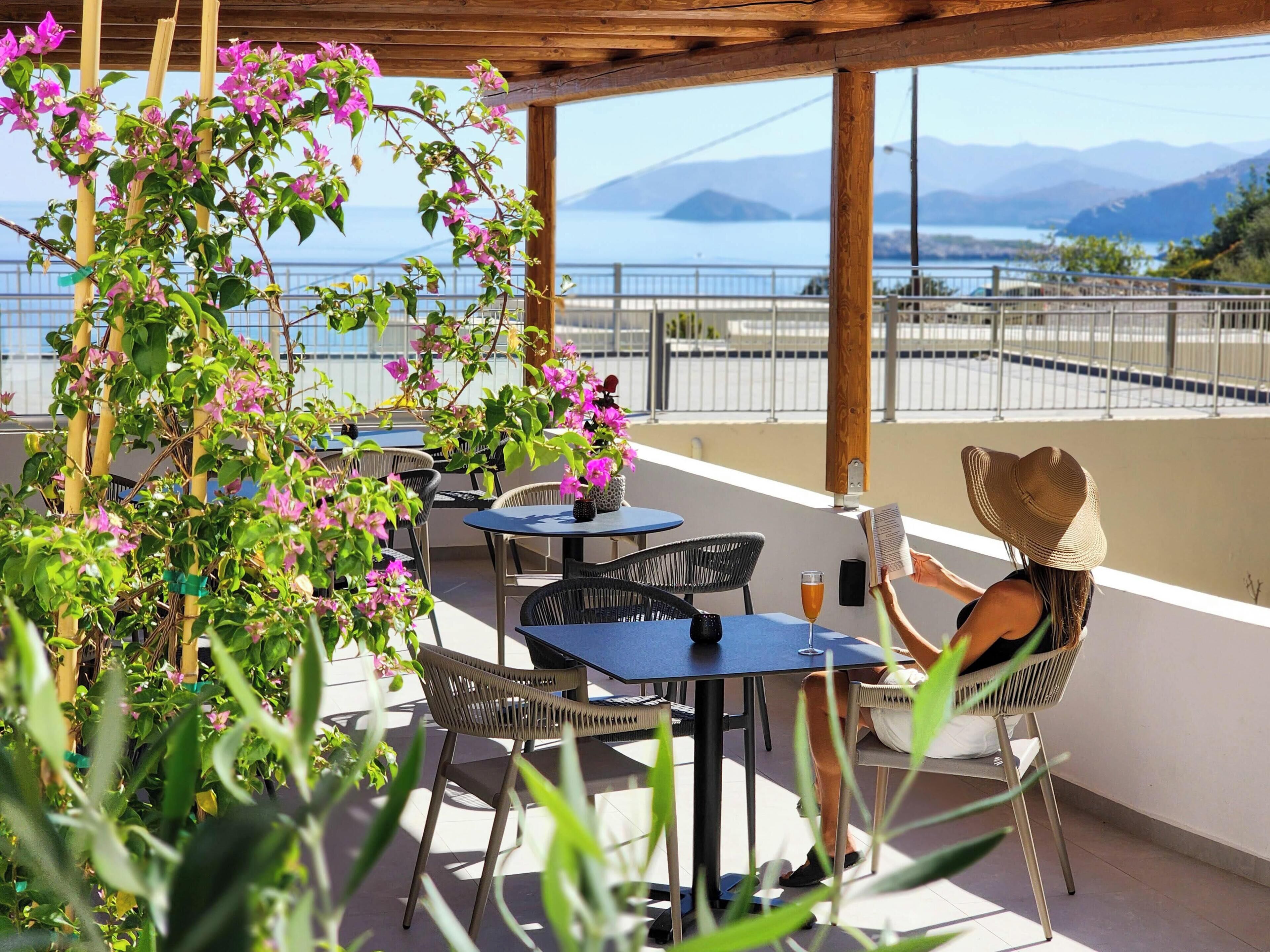 An outdoor terrace with tables and chairs, a pergola with flowering plants, and a view of the sea and mountains.