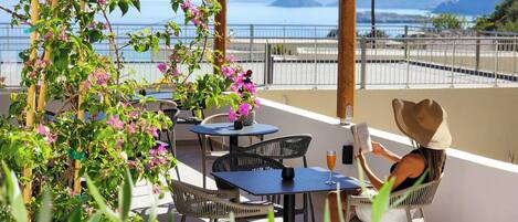 An outdoor terrace with tables and chairs, a pergola with flowering plants, and a view of the sea and mountains.
