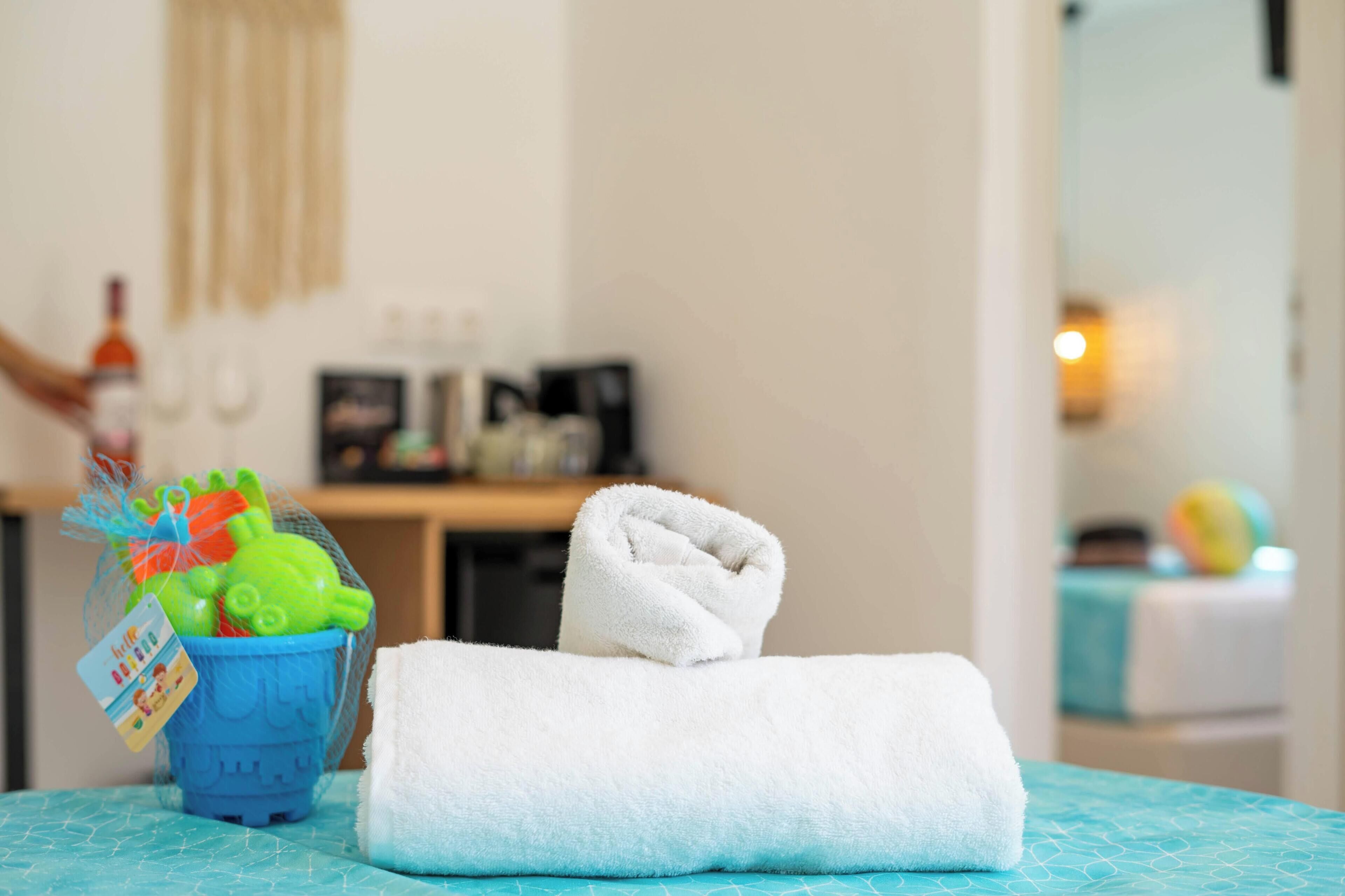 A neatly made bed with a white towel, a blue bucket with toys, and a bottle of water.