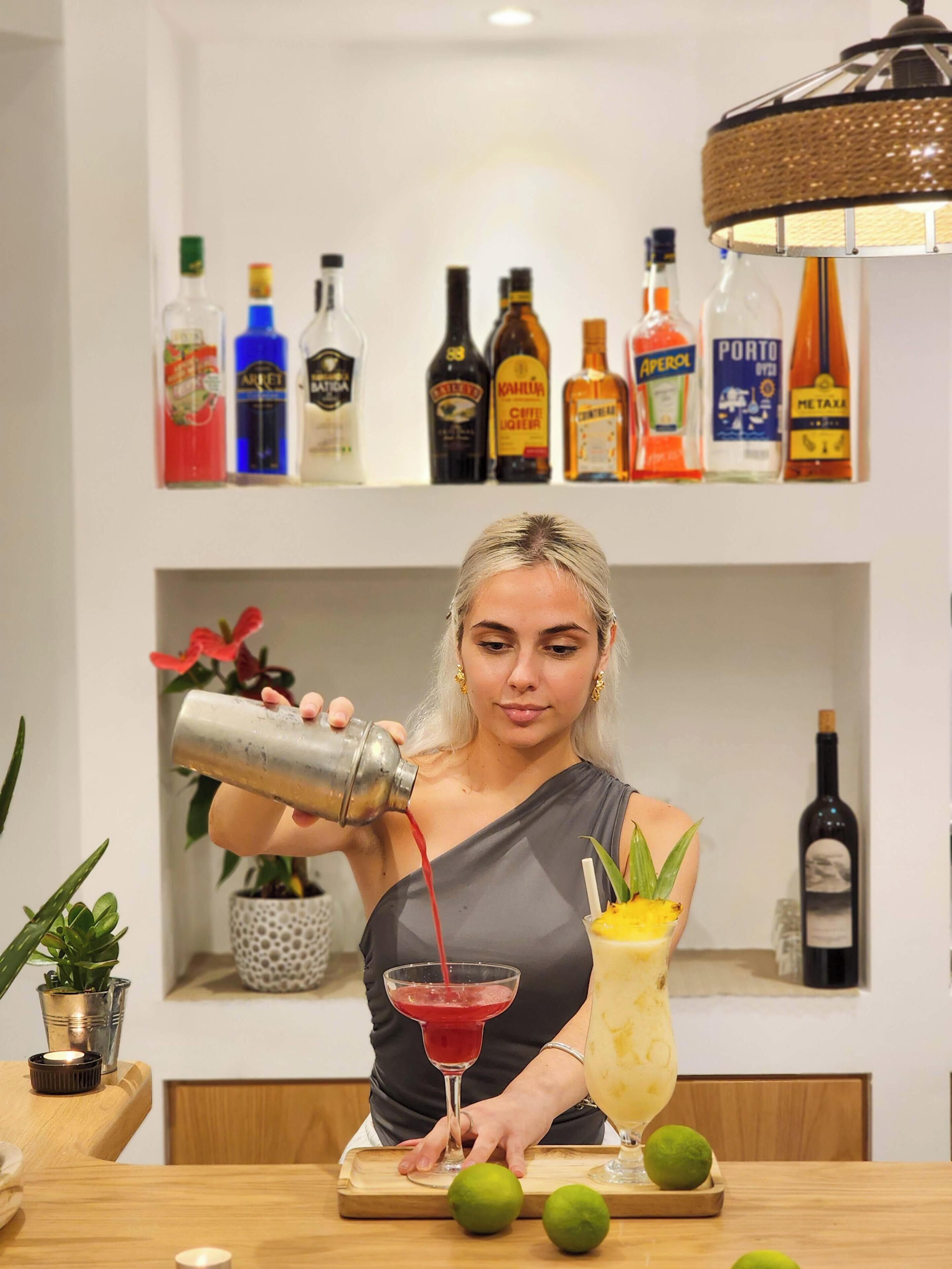 A person pouring a drink into a glass, with a variety of bottles on a shelf behind them.