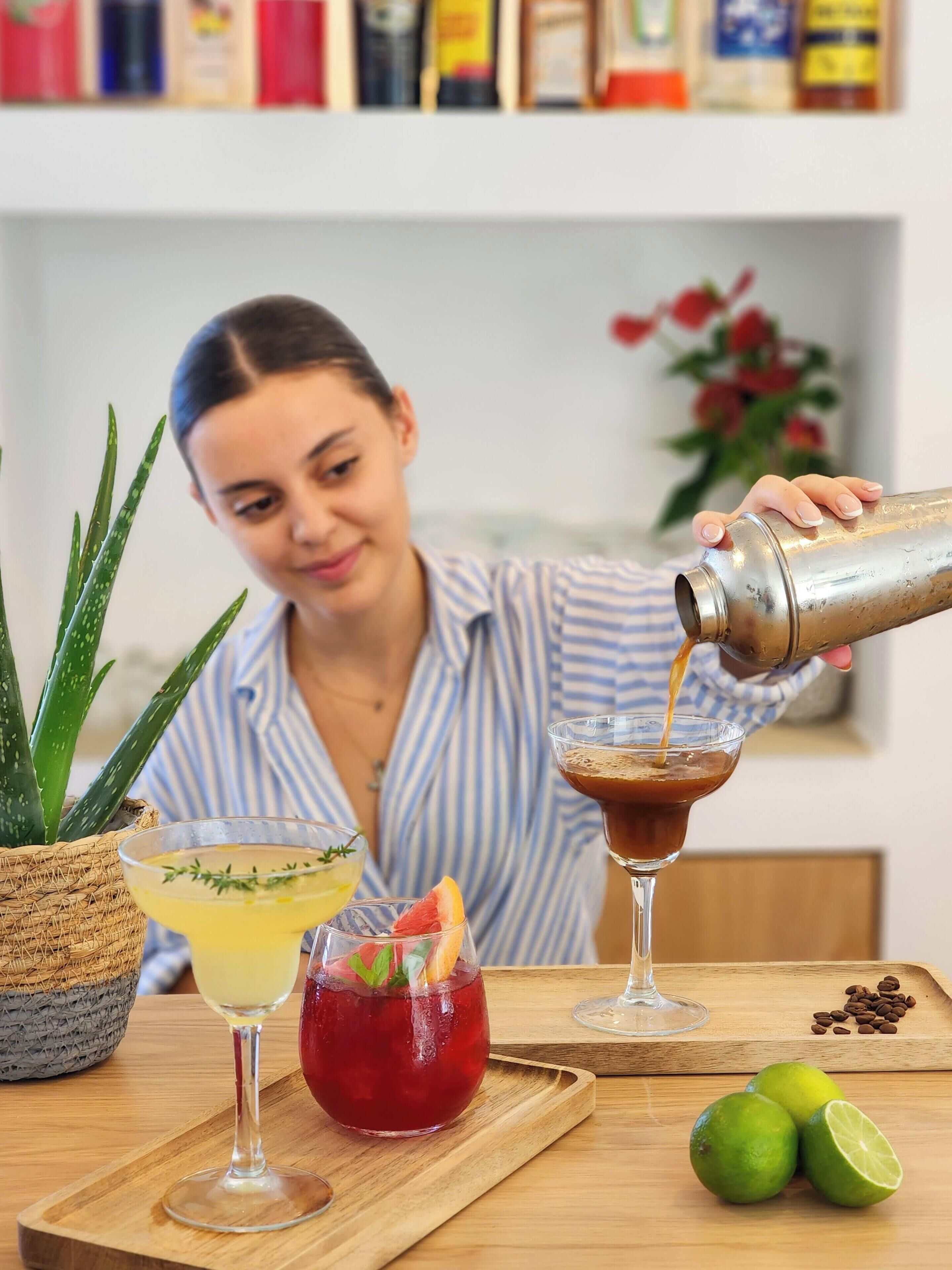 Una persona versa una bevanda in un bicchiere, con un tavolo di fronte. Ci sono due bicchieri, uno con una bevanda gialla e l'altro con una bevanda rossa, e una pianta sullo sfondo.