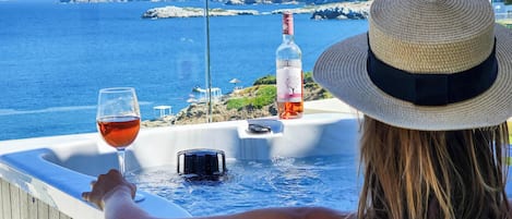 Una persona relajándose junto a una ventana con vistas al mar, una botella de vino y una copa de vino sobre una mesa.