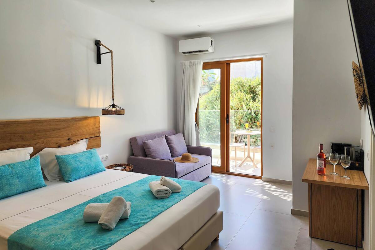 Une chambre d’hôtel avec un lit, un canapé, une petite table avec des verres et une bouteille, et une porte-fenêtre coulissante donnant sur l’extérieur.