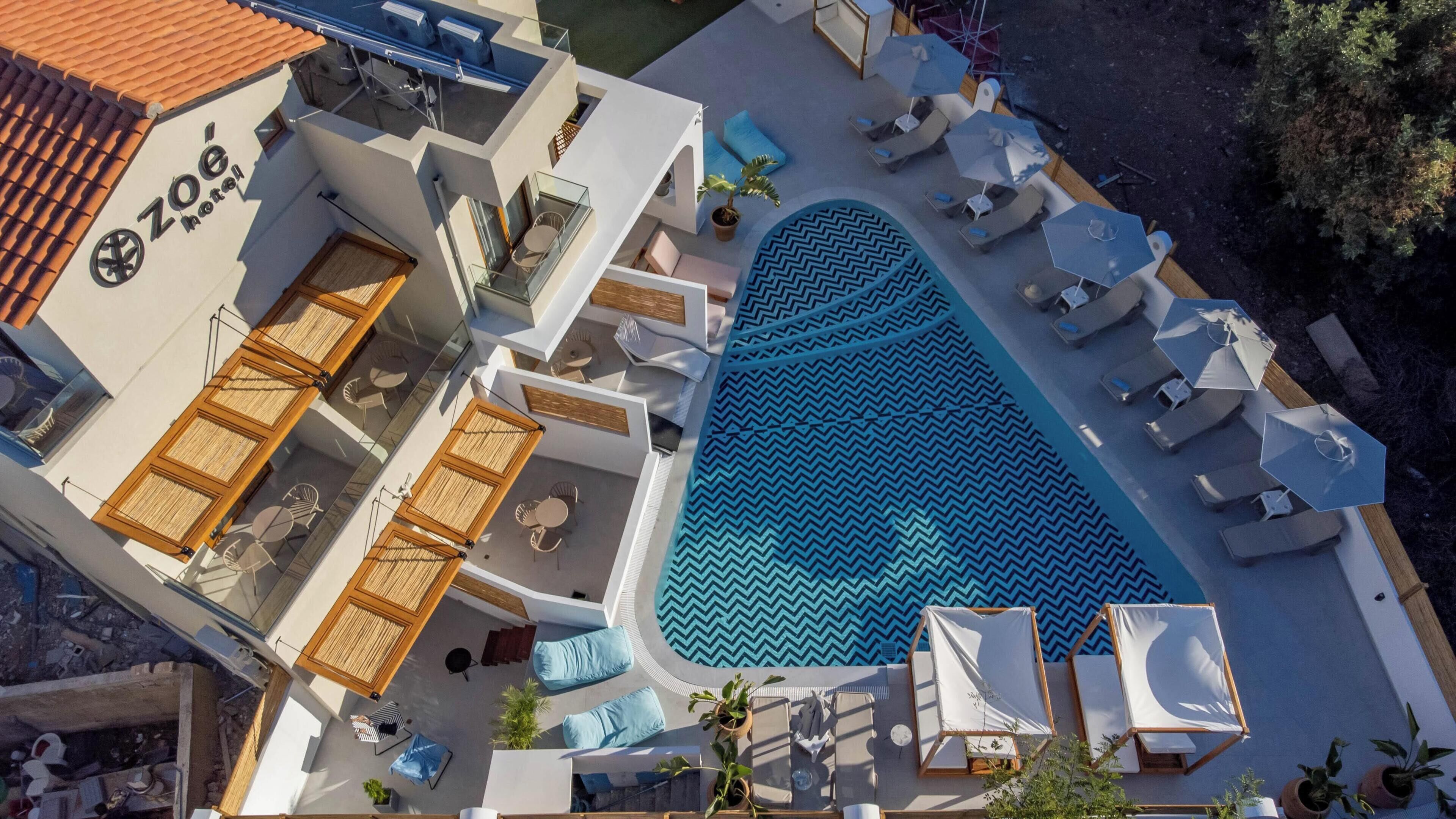 A rooftop pool area with lounge chairs, a pool with a chevron pattern, and a building with a sign reading 'Zoe Hotel'.