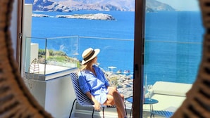 Eine Person sitzt auf einem Stuhl neben einer Glastür und blickt auf eine Küstenlandschaft mit Bergen und dem Meer.