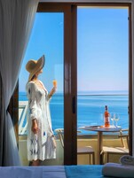 Una habitación con balcón que da al mar, una mesa con una botella y copas, y una cama con una toalla enrollada.