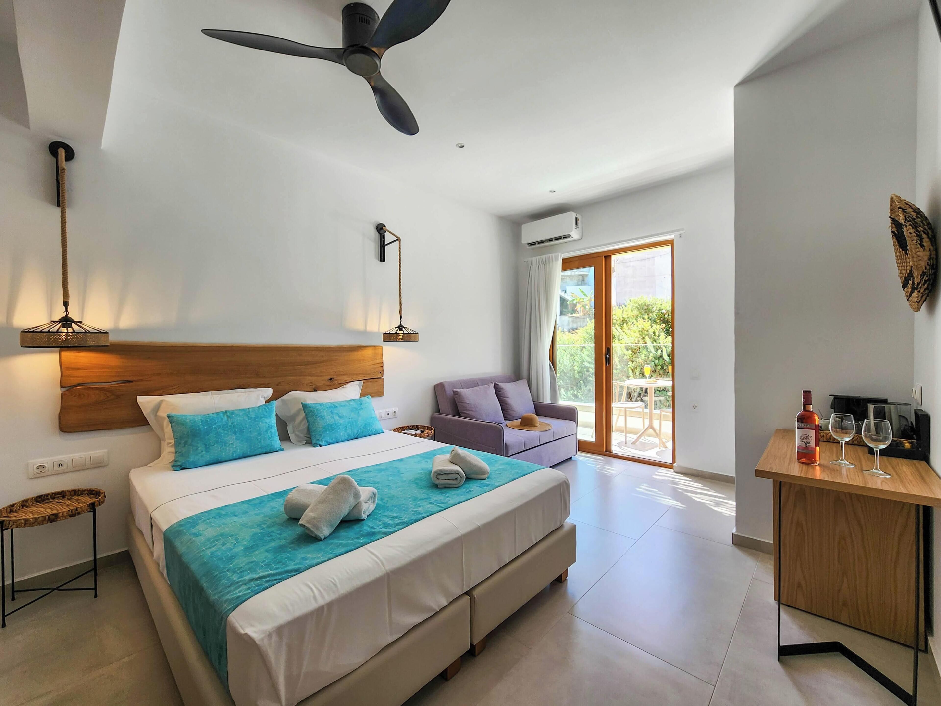 Une chambre à coucher avec un lit, un ventilateur de plafond, un canapé, une table avec une bouteille et des verres, et une porte-fenêtre coulissante donnant sur l’extérieur.