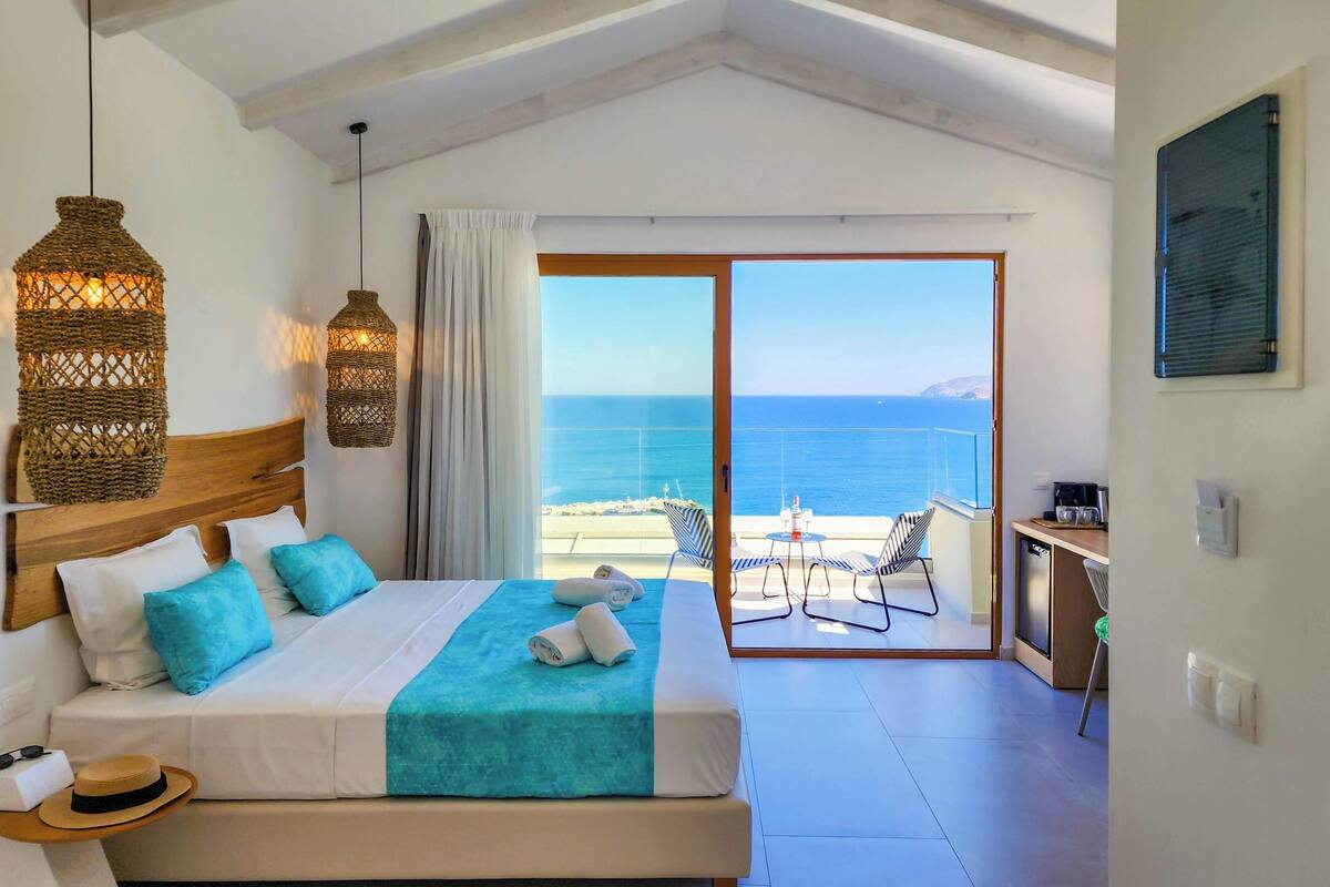 Une chambre avec un lit, une vue sur la mer, un balcon avec des chaises et une petite kitchenette.