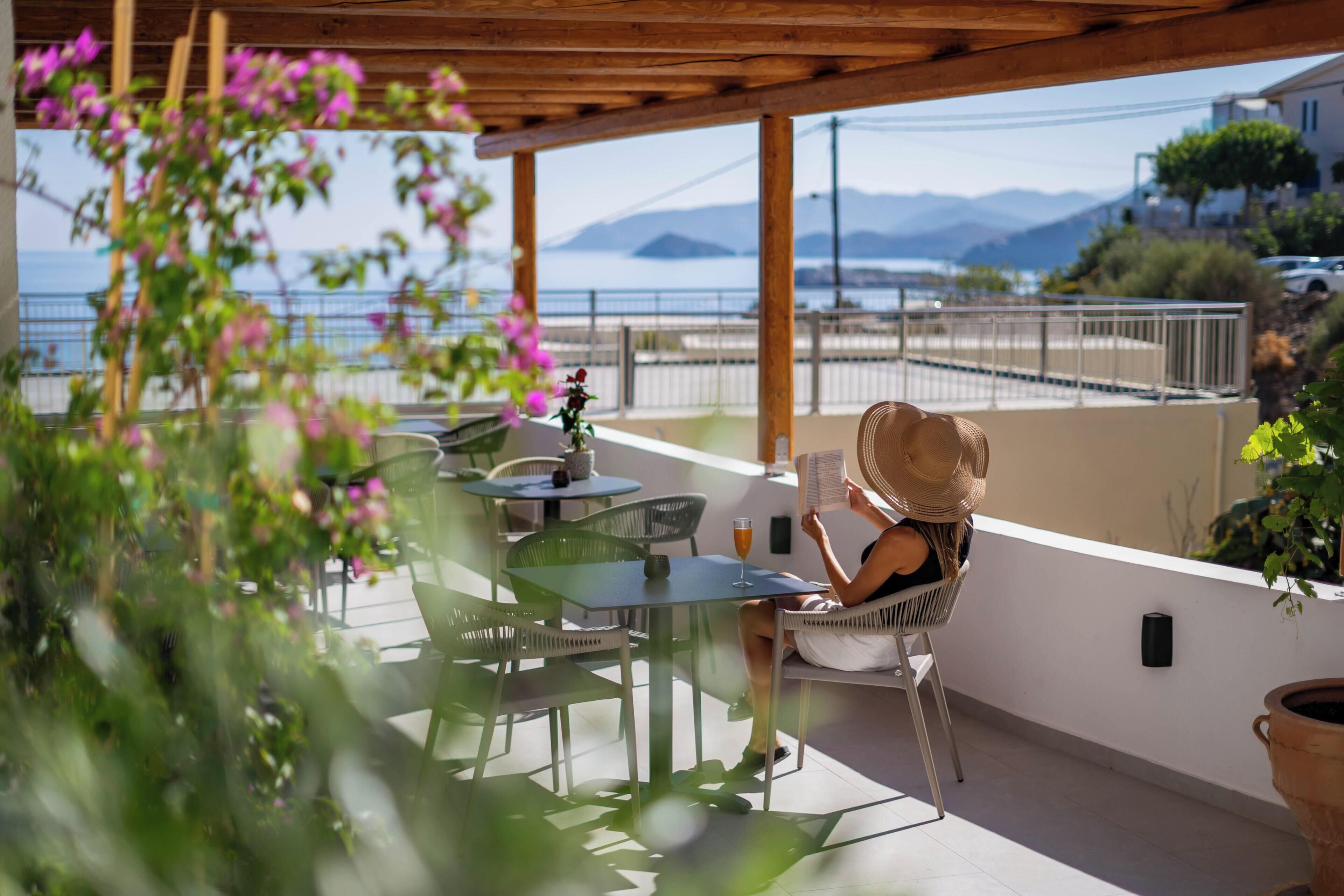 A terrace with a view of the sea, a table with chairs, and a person reading a book.