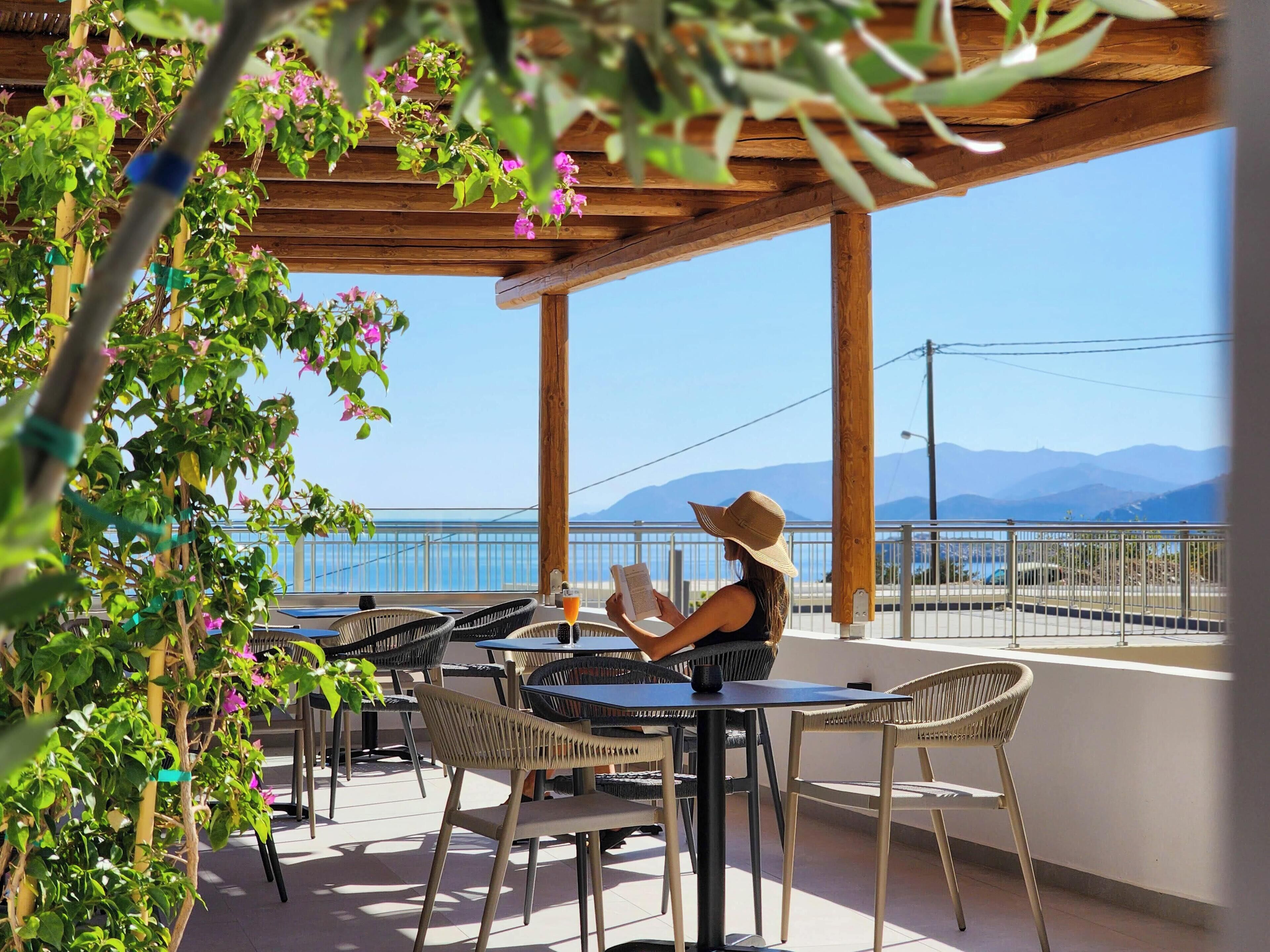 A covered outdoor seating area with wicker chairs and tables, overlooking a body of water and mountains.