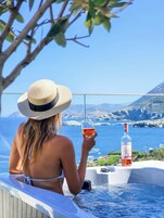 Una persona con traje de baño blanco y sombrero de paja sentada junto a un jacuzzi, con una botella de vino y una copa de vino tinto, con vistas a un cuerpo de agua y montañas.