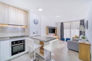 A modern kitchen with a built-in oven, a central island with bar stools, a grey sofa, and a TV in the living area.