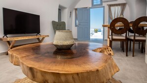 A wooden dining table with a bowl centerpiece, a television, and a chair in a room with a view through an open door. - Luxury Cave Suite | Jetted Tub | Sea & Sunset View (Oía)