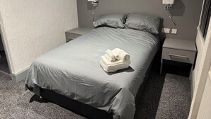 Un lit bien fait, avec une couette et des oreillers gris, une petite table de chevet avec une lampe et une prise électrique fixée au mur.