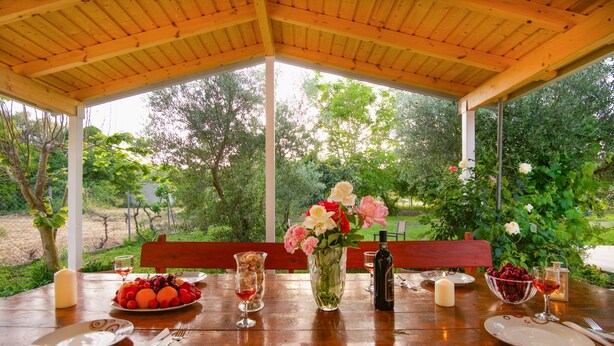 Une table en bois installée à l’extérieur, mise avec des assiettes, des couverts, un vase de fleurs et un saladier de fruits.