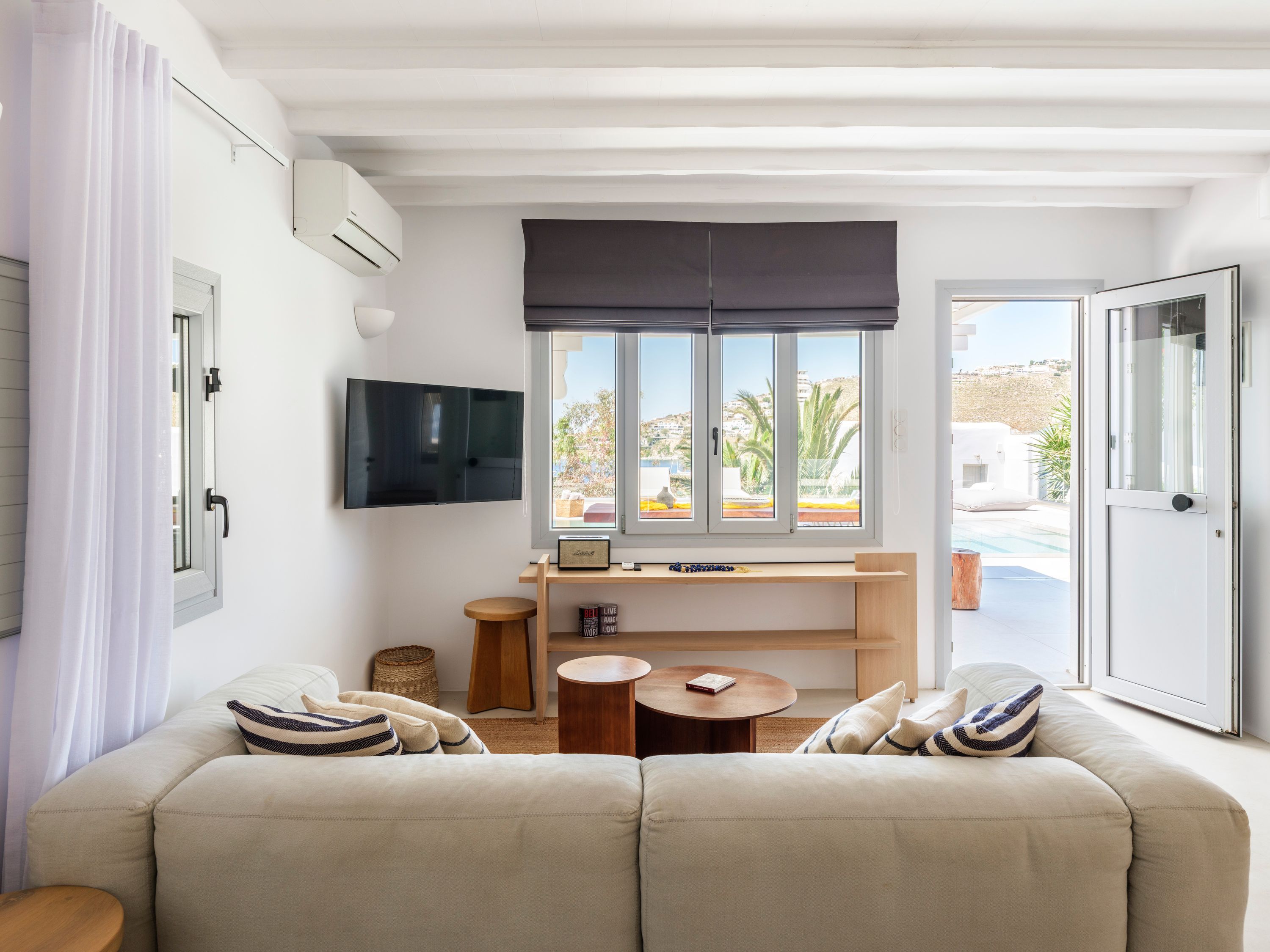 Uma sala de estar moderna com um sofá, uma pequena mesa de madeira, uma TV fixada na parede e uma janela com vista para uma piscina e edifícios.