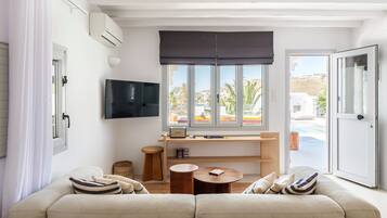 Uma sala de estar moderna com um sofá, uma pequena mesa de madeira, uma TV fixada na parede e uma janela com vista para uma piscina e edifícios.