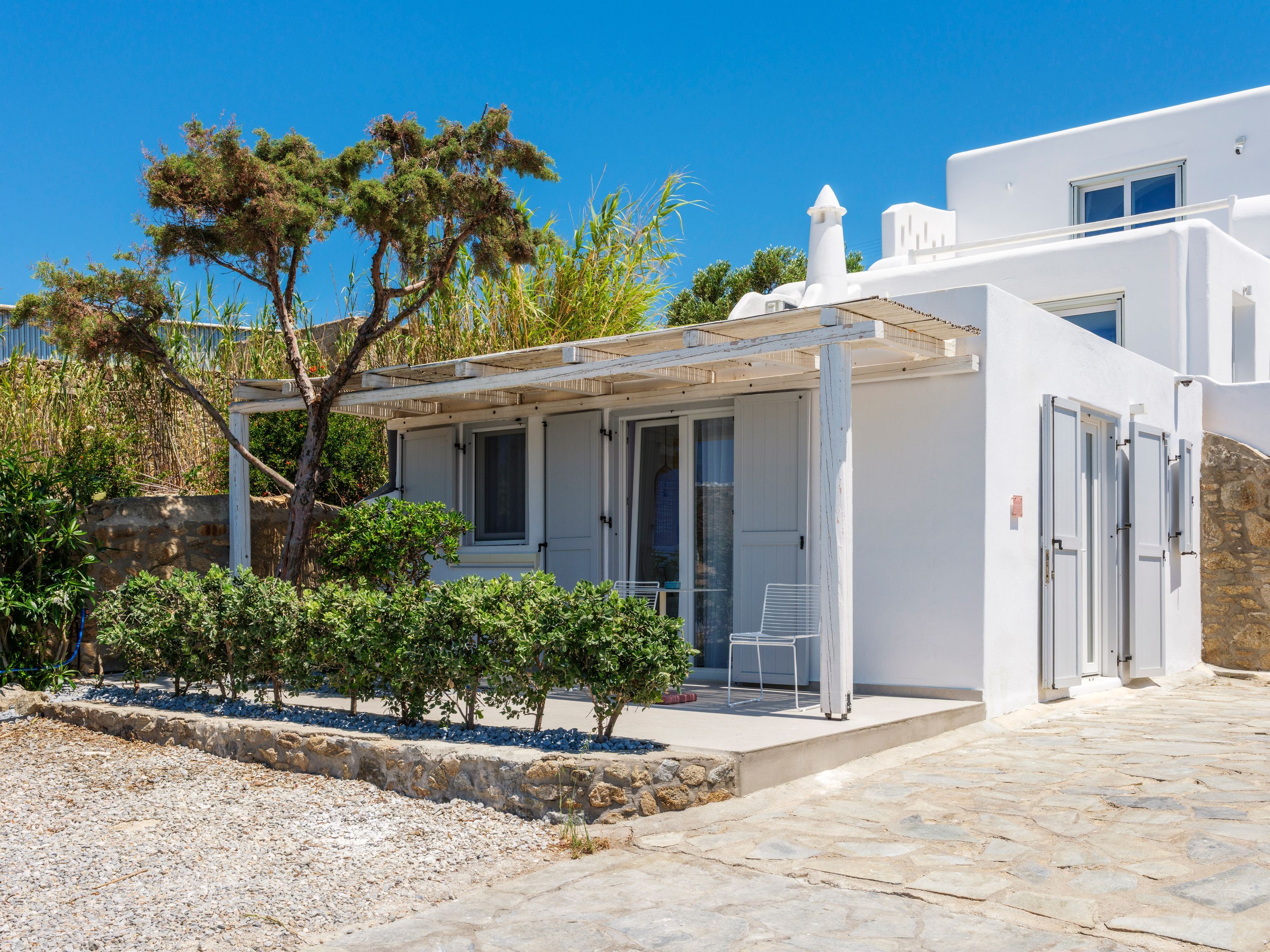 A white building with a covered patio, stone flooring, and a small garden area with shrubs and a tree.