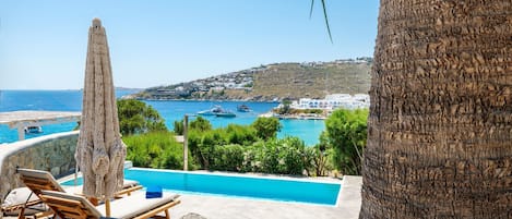 Zona de piscina con sillones de descanso y una sombrilla, con vistas a un pueblo costero y al mar.