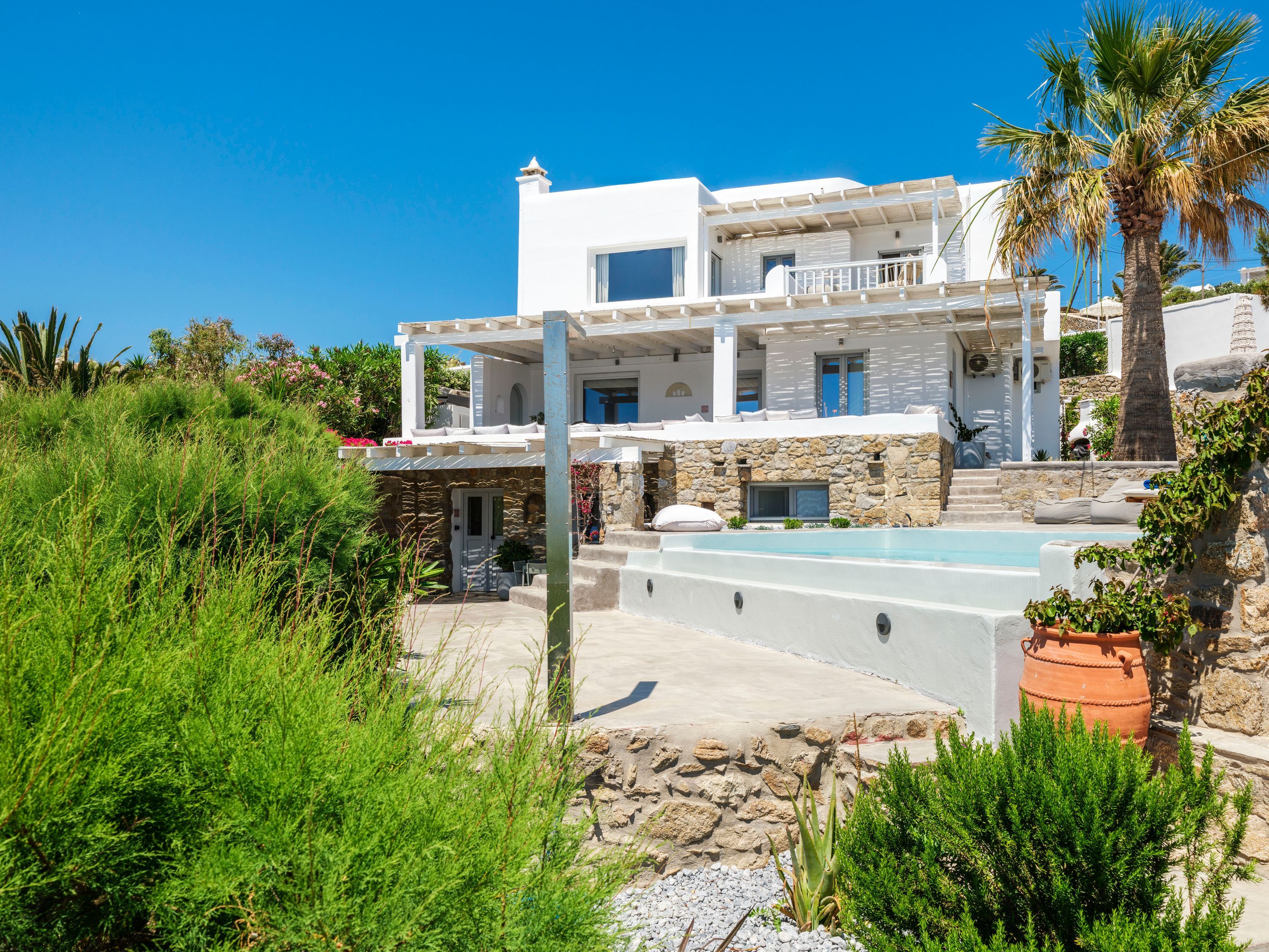Eine moderne weiße Villa mit Pool, umgeben von viel Grün und blauem Himmel.