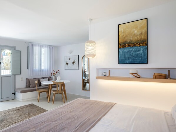 A modern hotel room with a bed, a dining area with a table and chairs, a sofa, and a large window with curtains. - myMykonos Guest House (Platis Gialos)
