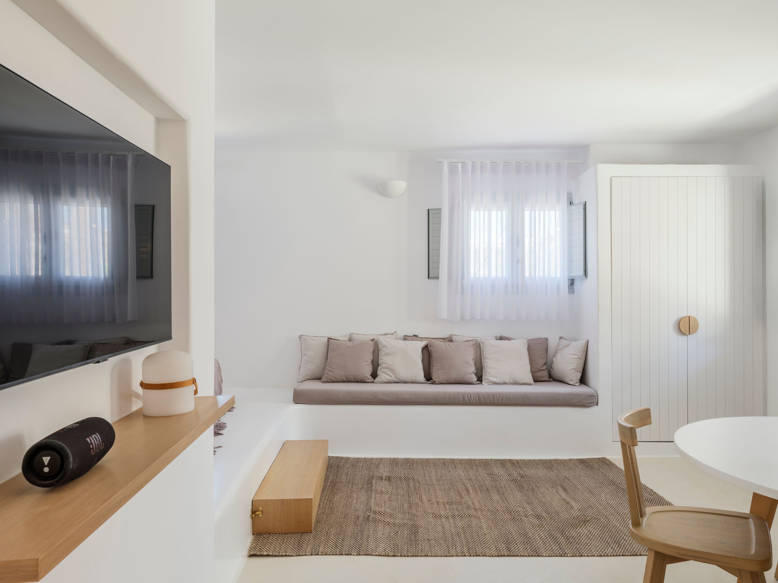 A modern living room with a flat-screen TV, a sofa with cushions, a wooden side table, a round table with chairs, and a woven rug.