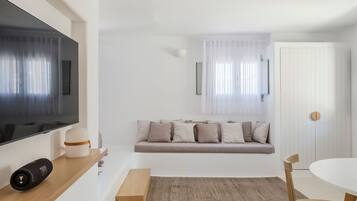 A modern living room with a flat-screen TV, a sofa with cushions, a wooden side table, a round table with chairs, and a woven rug.