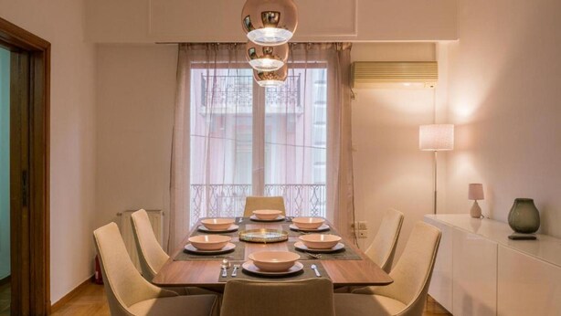 En matplats med ett träbord dukat för sex personer, sex beige stolar, en kristallkrona och ett fönster med tunna, genomskinliga gardiner.
