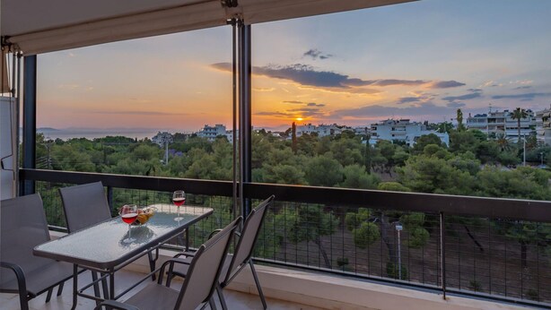 A balcony with a table and chairs, overlooking a cityscape with buildings and trees during sunset. - Archer - Sea view Apartment (Voula)