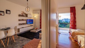 A modern hotel room with a dining area, a TV, a balcony with red curtains, and a view of the city. - Archer - Sea view Apartment (Voula)