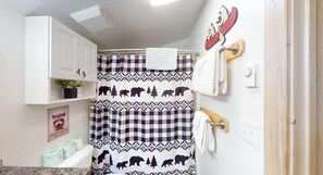 A bathroom with a checkered shower curtain, a mounted towel rack, and a shelf with a plant. - Pet-Friendly | Yellowstone 31 MI | Bear Cave 2 (Island Park)