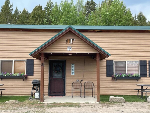 A wooden building with a green roof, a welcome sign, and two metal chairs outside.