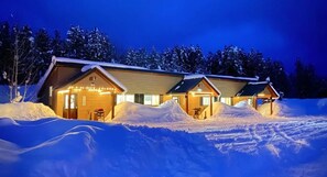A row of log cabins with snow-covered roofs, surrounded by a snowy landscape at night.