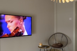 A television mounted on a wall, a round chair with a cushion, a small table with a candle and a book, and a modern light fixture. - Demetra - Koukaki center apartment (Athina)