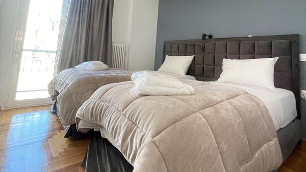 Un dormitorio con dos camas, un cabecero gris, una lámpara colgante y una ventana con cortinas.