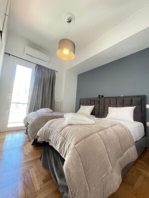 A bedroom with two beds, a grey headboard, a hanging light fixture, and a window with curtains. - Plato - Acropolis Residence Apartment (Athina)