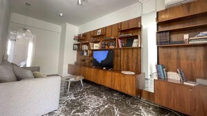 Una sala de estar moderna con un televisor de pantalla plana grande, estanterías de madera con libros, piso estampado en mármol y un sofá gris.