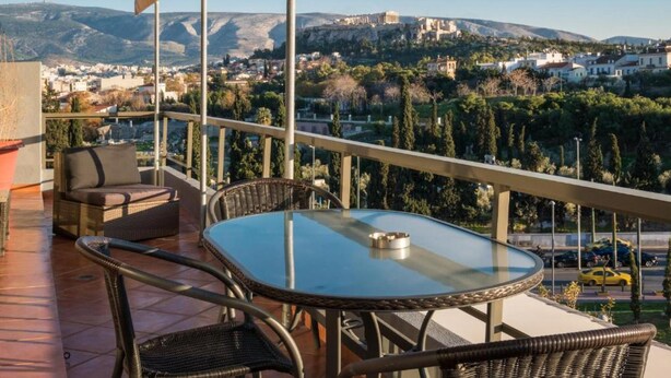 A balcony with wicker furniture, a glass table, and a view of a cityscape and mountains. - Virgo - Loft with Spectacular View to Acropolis (Athina)