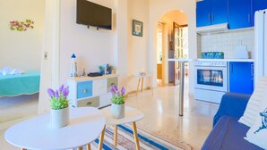 Ein modernes Wohnzimmer mit einer Couch, einem Esstisch mit zwei Blumentöpfen, einer Küche mit blauen Schränken und einem weißen Kühlschrank sowie einem an der Wand montierten Flachbildfernseher.